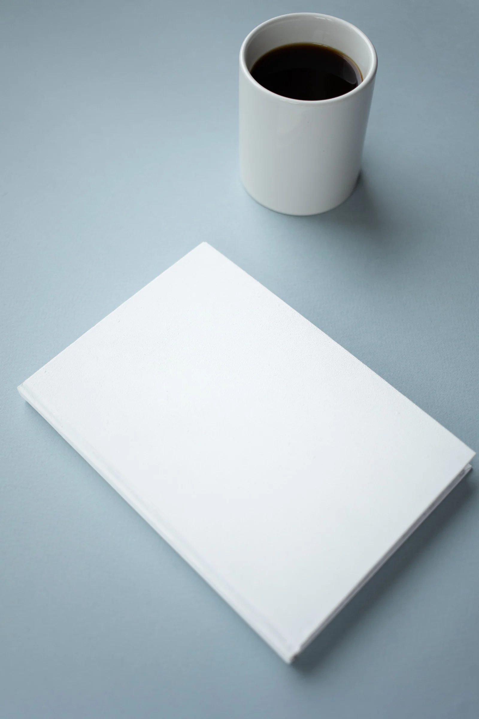 Cuaderno cerrado de tapa blanca junto a una taza de café sobre una mesa celeste.