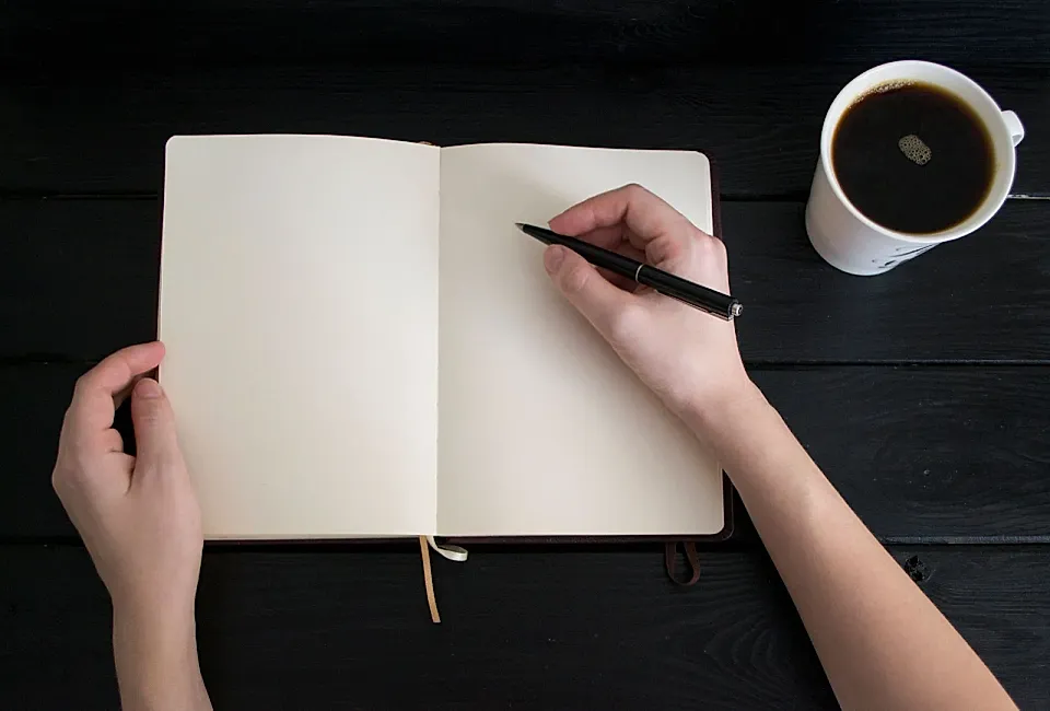 Manos con un bolígrafo sobre un cuaderno abierto junto a una taza de café.