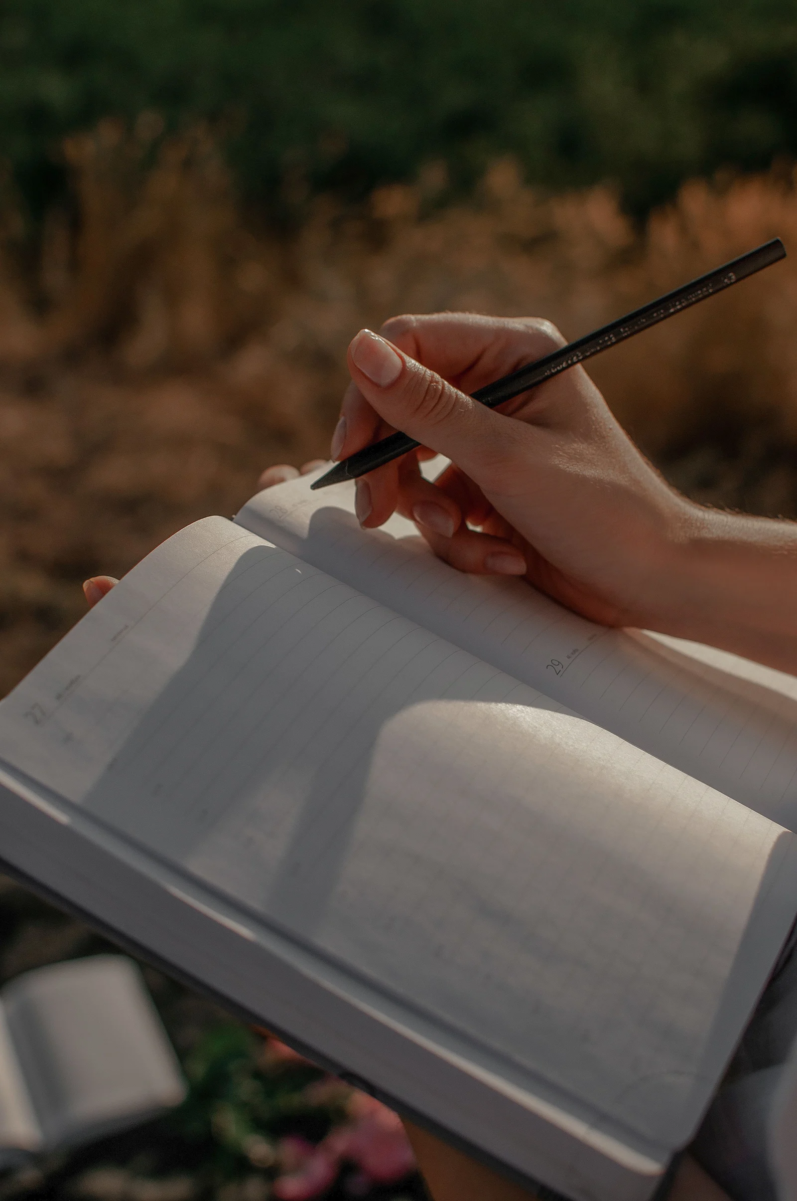 Una mano escribe con lápiz en una libreta rayada al aire libre.