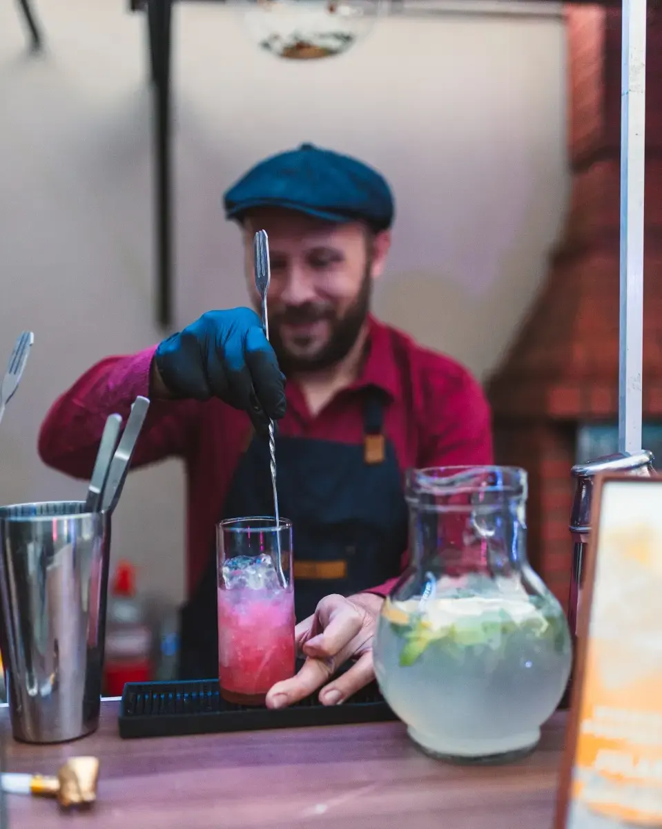 Bartender preparando un trago rosado junto a las herramientas y bebidas de la barra